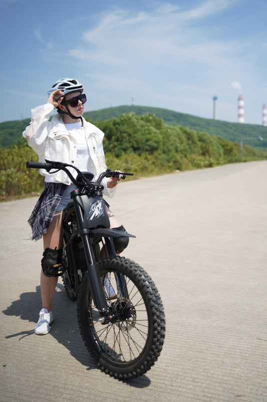 Woman wearing helmet and protective gear standing next to electric bike on open road with green hills in background